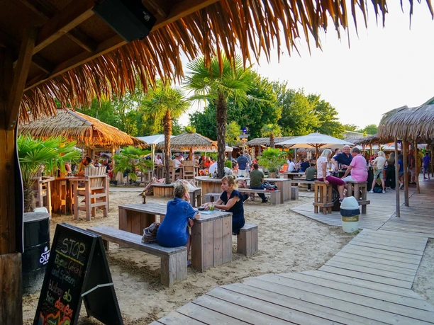 Aloha-Beachclub Herford, L Schneider (2).jpeg Strandbar mit Holzmöbeln und Palmen, Menschen genießen Getränke bei Sonnenuntergang in tropischem Flair.