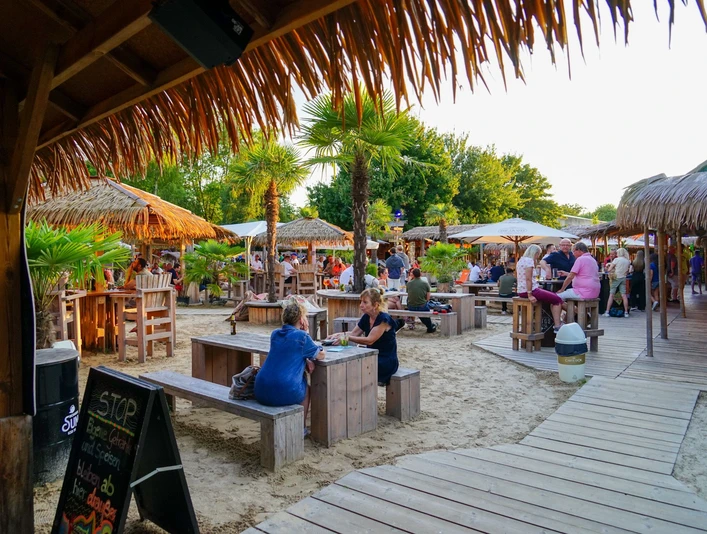 Aloha-Beachclub Herford, L Schneider (2).jpeg Strandbar mit Holzmöbeln und Palmen, Menschen genießen Getränke bei Sonnenuntergang in tropischem Flair.