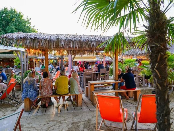 Menschen entspannen auf Holzsitzecken unter Palmen mit Strohschirmen, umgeben von sandigem Boden.