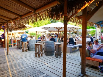 Aloha-Beachclub Herford, L Schneider (4).jpeg Offene, luftige Strandbar mit Holzboden, Palmdach und Sitzgelegenheiten unter Sonnenschirmen.