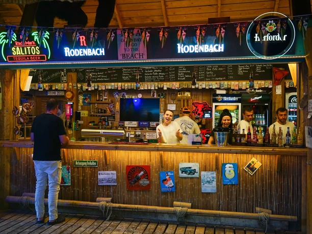 Eine offene Strandbar mit hölzernem Tresen im Aloha-Beachclub, betrieben von freundlichem Personal.
