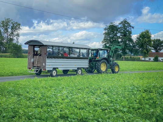 Planwagenfahrt.jpg Ein Traktor zieht einen Planwagen über eine ländliche Straße, umgeben von grünen Feldern.