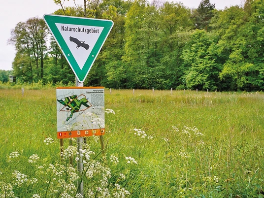 Salzenteichs_Heide.jpg Schild "Naturschutzgebiet" mit Adler-Symbol auf einer Wiese vor einem bewaldeten Hintergrund.