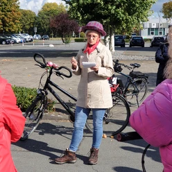 20221008_135734.jpg Eine Frau in beiger Jacke und Hut dirigiert eine Radgruppe auf einem sonnigen Parkplatz.