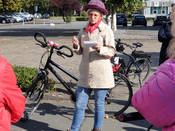20221008_135734.jpg Eine Frau in beiger Jacke und Hut dirigiert eine Radgruppe auf einem sonnigen Parkplatz.