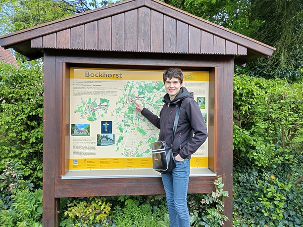 IMG_20240416_180536_HDR.jpg Ein junger Mann, der vor einer großen Infotafel in Bockhorst im Freien lächelt und posiert.