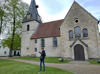 IMG_20240416_181117_HDR.jpg Historische Kirche aus Stein mit Satteldach und Glockenturm, im Vordergrund eine Person auf dem Rasen.