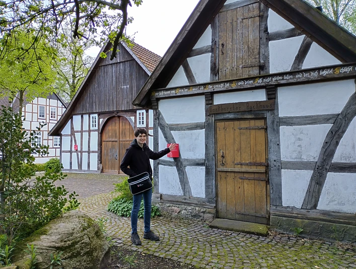 IMG_20240416_180329_HDR.jpg Historische Fachwerkhäuser in einem grünen Freilichtmuseum, Person hält eine rote Tasche.