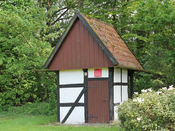 IMG_0245.JPG Fachwerkhäuschen in grünem Park, umgeben von Büschen und Bäumen, mit rotem Schild an der Wand.