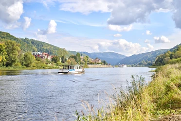 A river with a small boat in the foreground, surrounded by green hills and a village with red roofs under a blue sky with clouds.