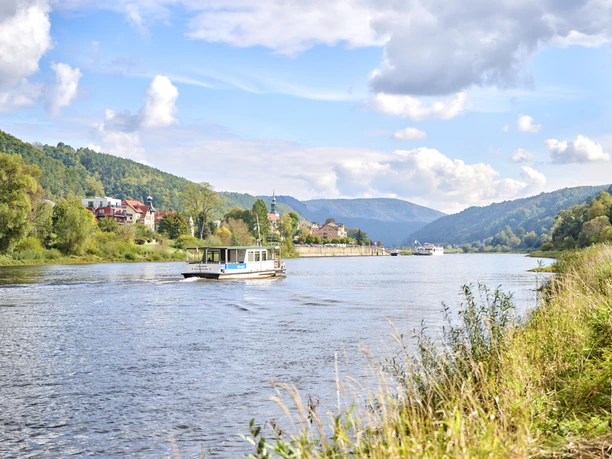 Bad Schandau Elbfähre Ein Fluss mit einem kleinen Boot im Vordergrund, umgeben von grünen Hügeln und einem Dorf mit roten Dächern unter einem blauen Himmel mit Wolken.