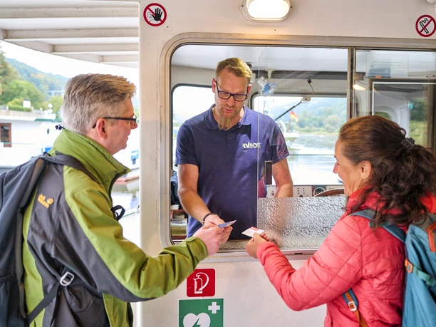Een man in een groene jas en een vrouw in een rode jas kopen kaartjes van een verkoper bij een botenloket; op de achtergrond is water te zien.