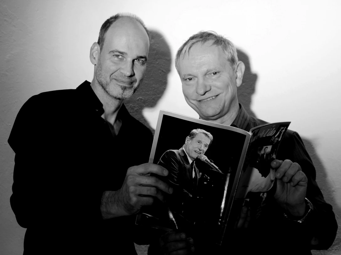 Pressefoto_UDO.jpg Zwei Männer, lächelnd, halten gemeinsam ein Buch in die Kamera. Der Hintergrund ist schlicht.