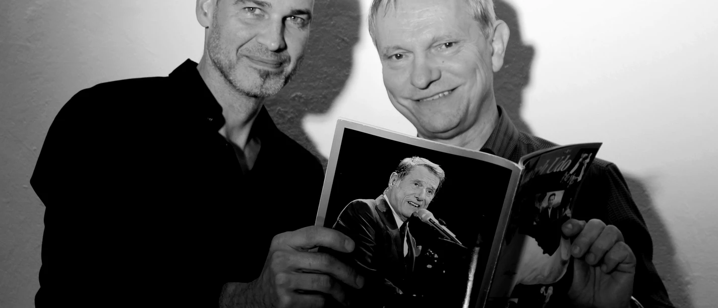 Pressefoto_UDO.jpg Zwei Männer, lächelnd, halten gemeinsam ein Buch in die Kamera. Der Hintergrund ist schlicht.