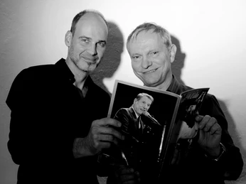 Pressefoto_UDO.jpg Zwei Männer, lächelnd, halten gemeinsam ein Buch in die Kamera. Der Hintergrund ist schlicht.