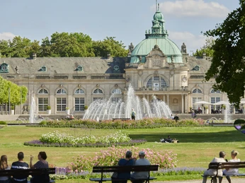 Kaiserpalais im Kurpark Barockes Gebäude mit Kuppel, Springbrunnen und Parkanlagen. Menschen auf Bänken genießen die Aussicht.