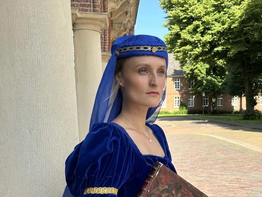 LAK: Almuth Cirksena Frau in königsblauem Mittelalterkleid mit Buch steht vor historischem Gebäude in Greetsiel.