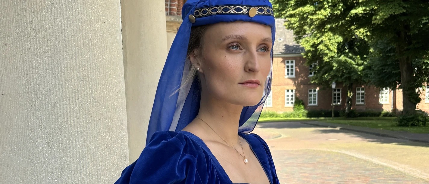 LAK: Almuth Cirksena Frau in königsblauem Mittelalterkleid mit Buch steht vor historischem Gebäude in Greetsiel.