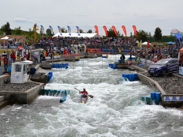99_Kanupark_Kanuslalom_KPM_b.JPG Kanufahrer navigiert durch die Stromschnellen eines künstlichen Kanals, gesäumt von Zuschauern.
