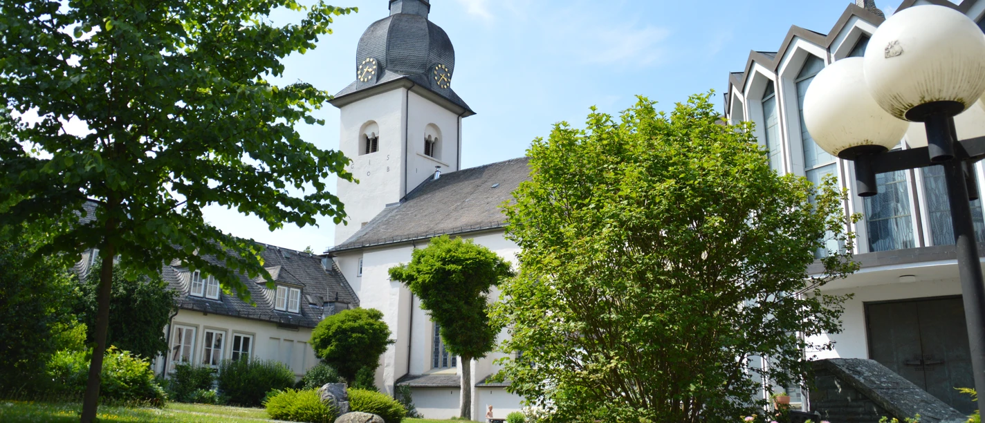 St. Walburga in Meschede mit weißem Kirchturm, Schieferdach, Bäumen und blauem Himmel.