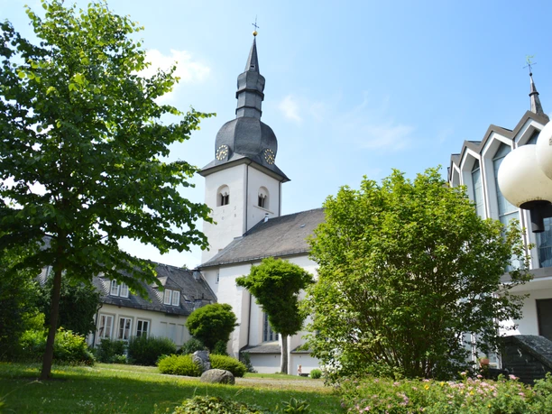 St. Walburga in Meschede mit weißem Kirchturm, Schieferdach, Bäumen und blauem Himmel.