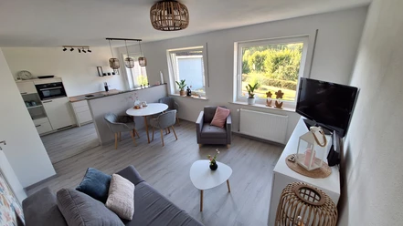 FeWo Diemelaue Bild 1.jpg Modernes Wohnzimmer mit grauer Couch, Esstisch und großer Fensterfront, die viel Licht hereinscheint.