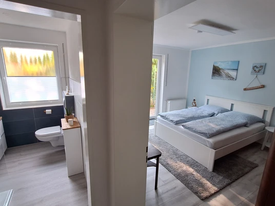 FeWo Diemelaue Bild 2.jpg Helles Schlafzimmer mit Doppelbett und Blick ins moderne Bad mit Fenster und grauen Fliesen.