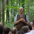 Ein älterer Mann steht im Wald und hält einen Zweig, umgeben von interessierten Zuhörern.