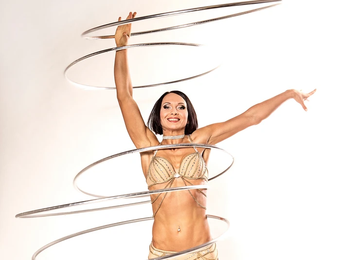 Olena 1.jpg Eine Frau in goldenen Outfits jongliert elegant acht Hula-Hoop-Reifen in einem Studio.