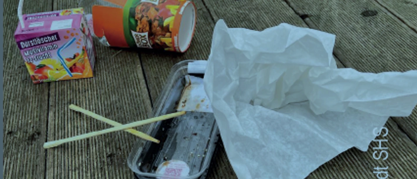 Verschiedene Abfälle, darunter Lebensmittelverpackungen und Plastikstraws, auf einem Holzboden verstreut.