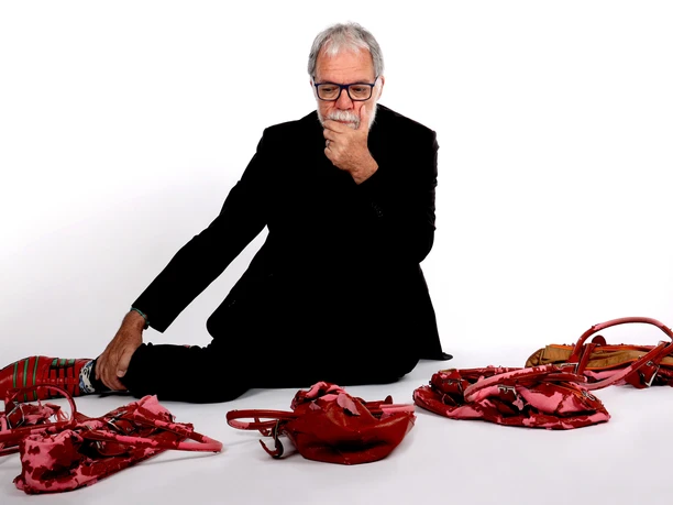 Wolfgang Trepper sitzt nachdenklich, umgeben von roten und rosa Handtaschen, auf einem weißen Boden.