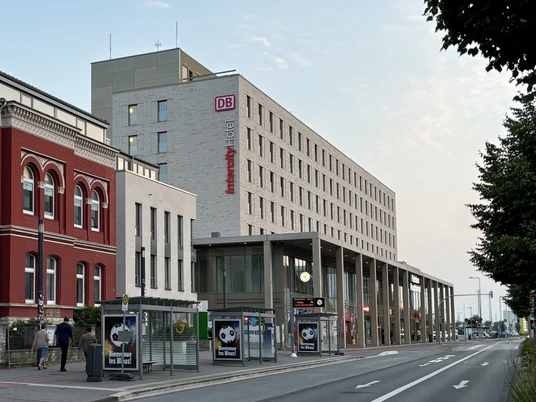 Paderborn Hauptbahnhof.jpg Paderborn Hauptbahnhof mit modernem Empfangsgebäude, Bahnhofsuhren und Werbeplakaten auf der Straße.