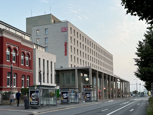 Paderborn Hauptbahnhof.jpg Paderborn Hauptbahnhof mit modernem Empfangsgebäude, Bahnhofsuhren und Werbeplakaten auf der Straße.