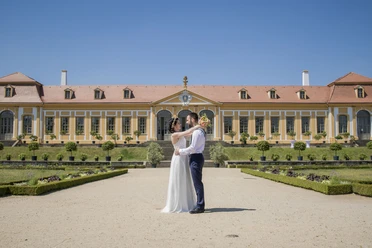 A wedding couple stands embracing in front of a historic building with a bright blue sky.