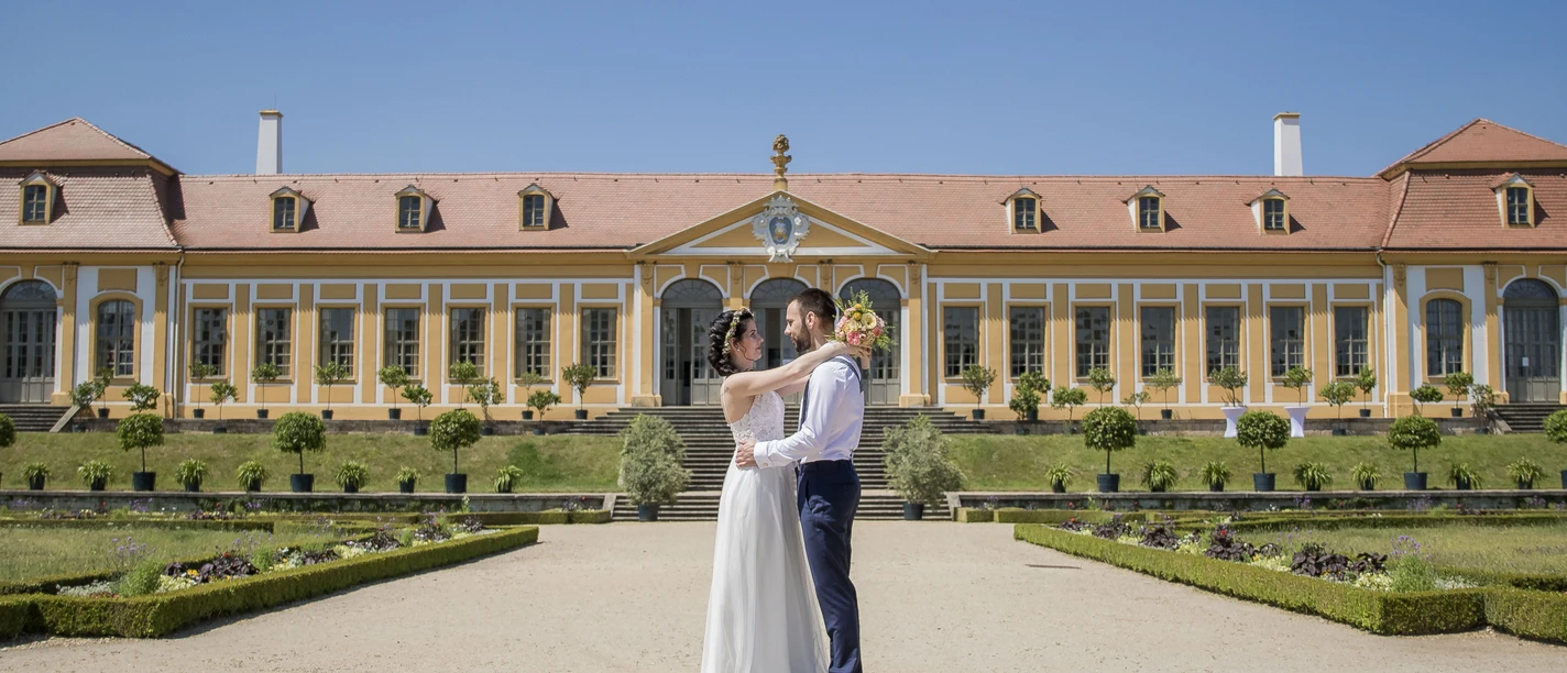 Ein Hochzeitspaar steht innig umarmt vor einem historischen Gebäude mit strahlend blauem Himmel.