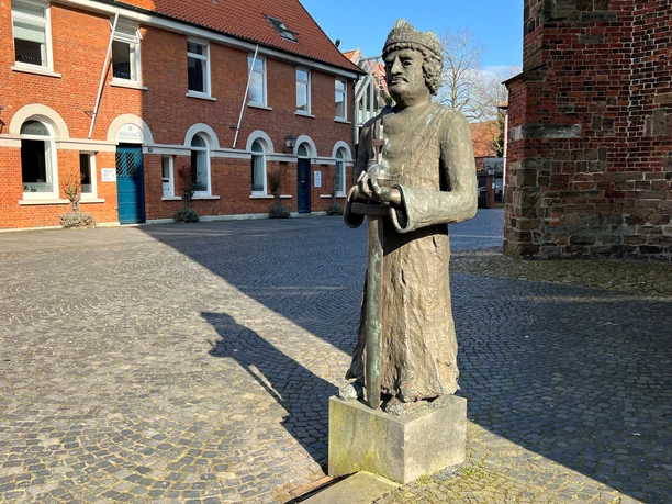 Eine bronzene Statue von Karl dem Großen steht auf einem gepflasterten Platz vor Backsteingebäuden.