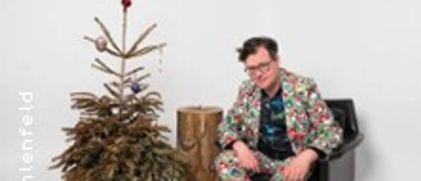 Ein Mann sitzt im gemusterten Anzug neben einem unterdekorierten Weihnachtsbaum auf einem schwarzen Stuhl.