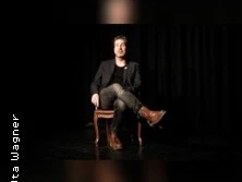 Ein Mensch sitzt in Lederstiefeln auf einem Stuhl vor einem dunklen Hintergrund.