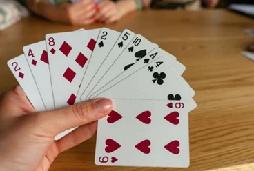 Canasta Eine Hand hält Karten mit Herzen, Karo, Pik und Kreuz. Zwei Personen spielen im Hintergrund.