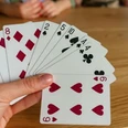 Canasta Eine Hand hält Karten mit Herzen, Karo, Pik und Kreuz. Zwei Personen spielen im Hintergrund.