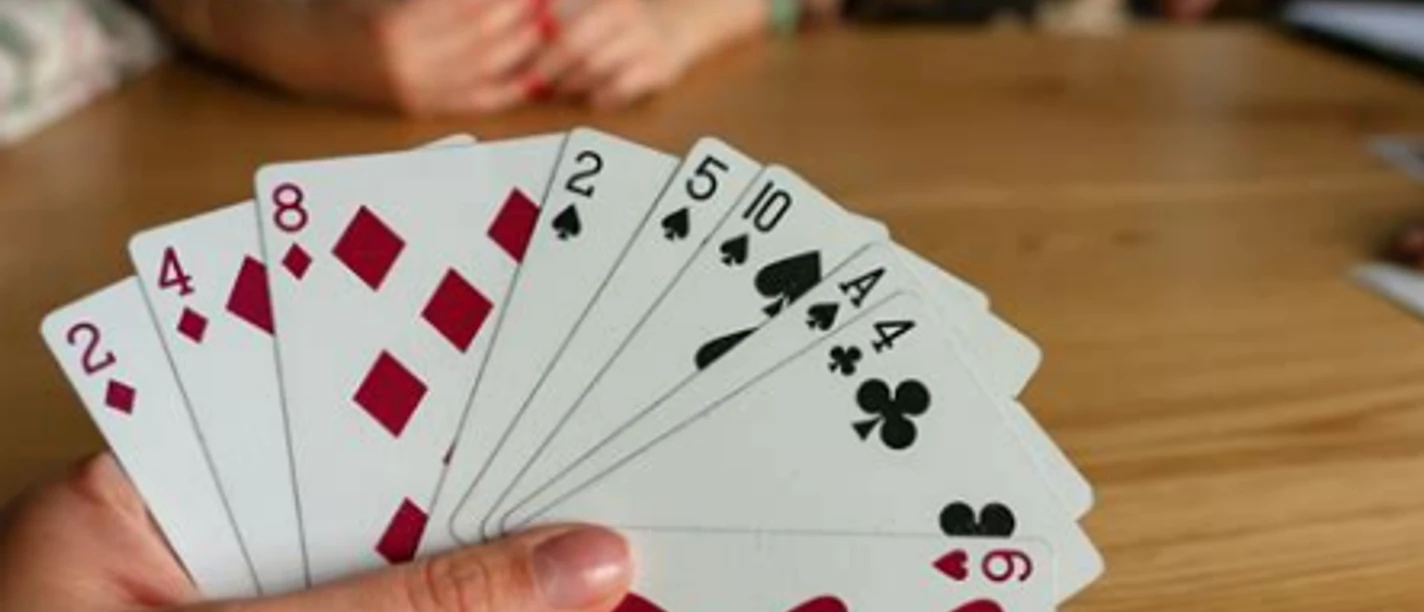 Canasta Eine Hand hält Karten mit Herzen, Karo, Pik und Kreuz. Zwei Personen spielen im Hintergrund.