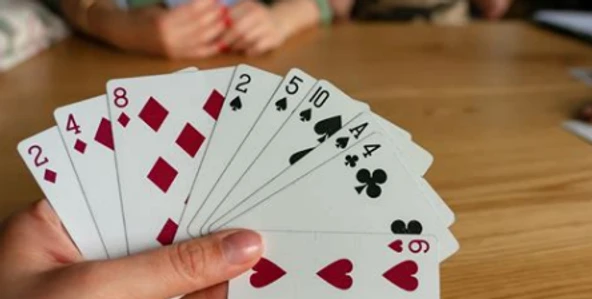 Canasta Eine Hand hält Karten mit Herzen, Karo, Pik und Kreuz. Zwei Personen spielen im Hintergrund.