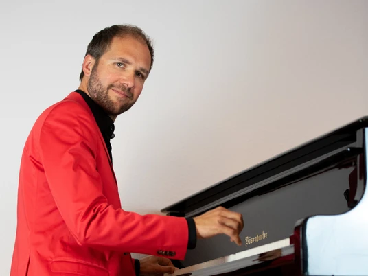 Stefan Ulbricht Stefan Ulbricht spielt im roten Anzug am Flügel und wirft dabei einen freundlichen Blick zur Seite.