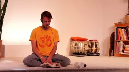 Yoga Ein Mann sitzt in entspannter Meditationshaltung auf einem Boden, trägt ein gelbes Om-Shirt und liest.