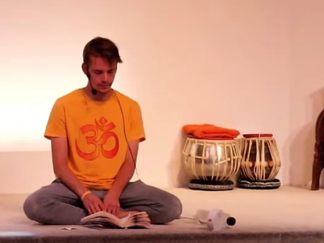 Yoga Ein Mann sitzt in entspannter Meditationshaltung auf einem Boden, trägt ein gelbes Om-Shirt und liest.