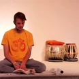 Yoga Ein Mann sitzt in entspannter Meditationshaltung auf einem Boden, trägt ein gelbes Om-Shirt und liest.