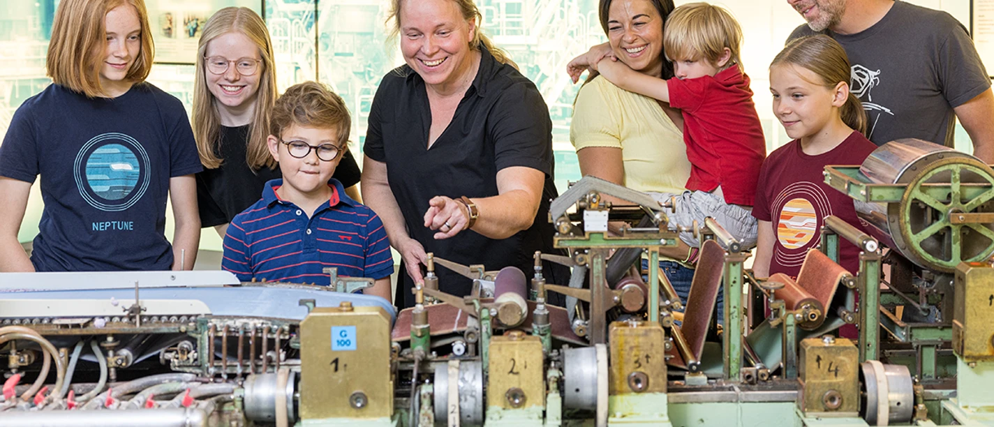 Kinderführung Laborpapiermaschine Eine Familie betrachtet interessiert eine historische Papiermaschine, geführt von einem Museumsmitarbeiter.