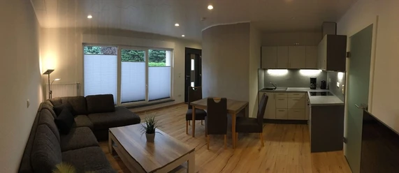 Bild 1.jpg Modern eingerichtetes Wohnzimmer mit grauem Sofa, Holzesstisch und offener Küchenzeile im Hintergrund.