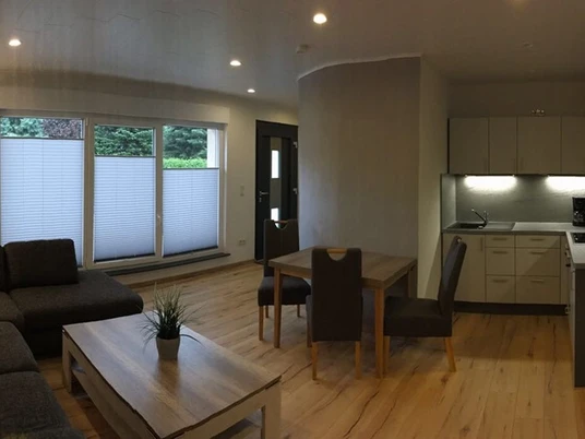 Bild 1.jpg Modern eingerichtetes Wohnzimmer mit grauem Sofa, Holzesstisch und offener Küchenzeile im Hintergrund.