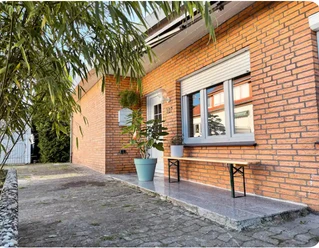 Fassade Senne Bungalow Backsteinfassade eines Hauses mit weißem Eingang, Fensterläden und Topfpflanze auf gepflastertem Weg.
